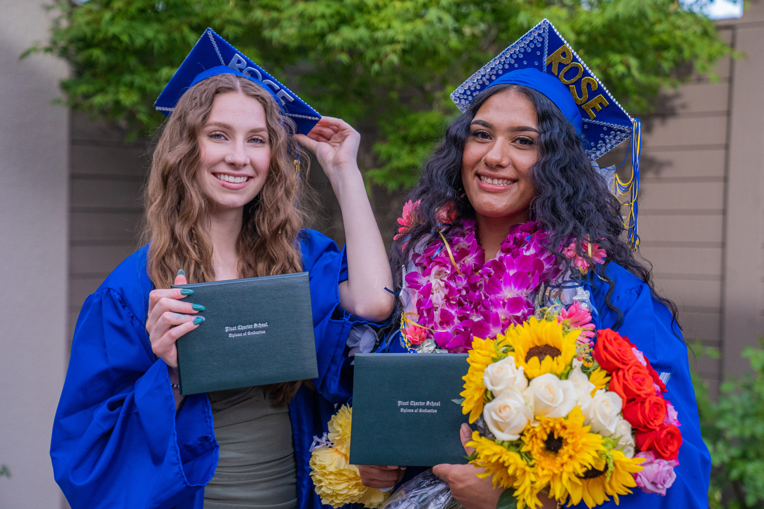 Two Pivot students wearing their graduation gowns and smiling with their diplomas